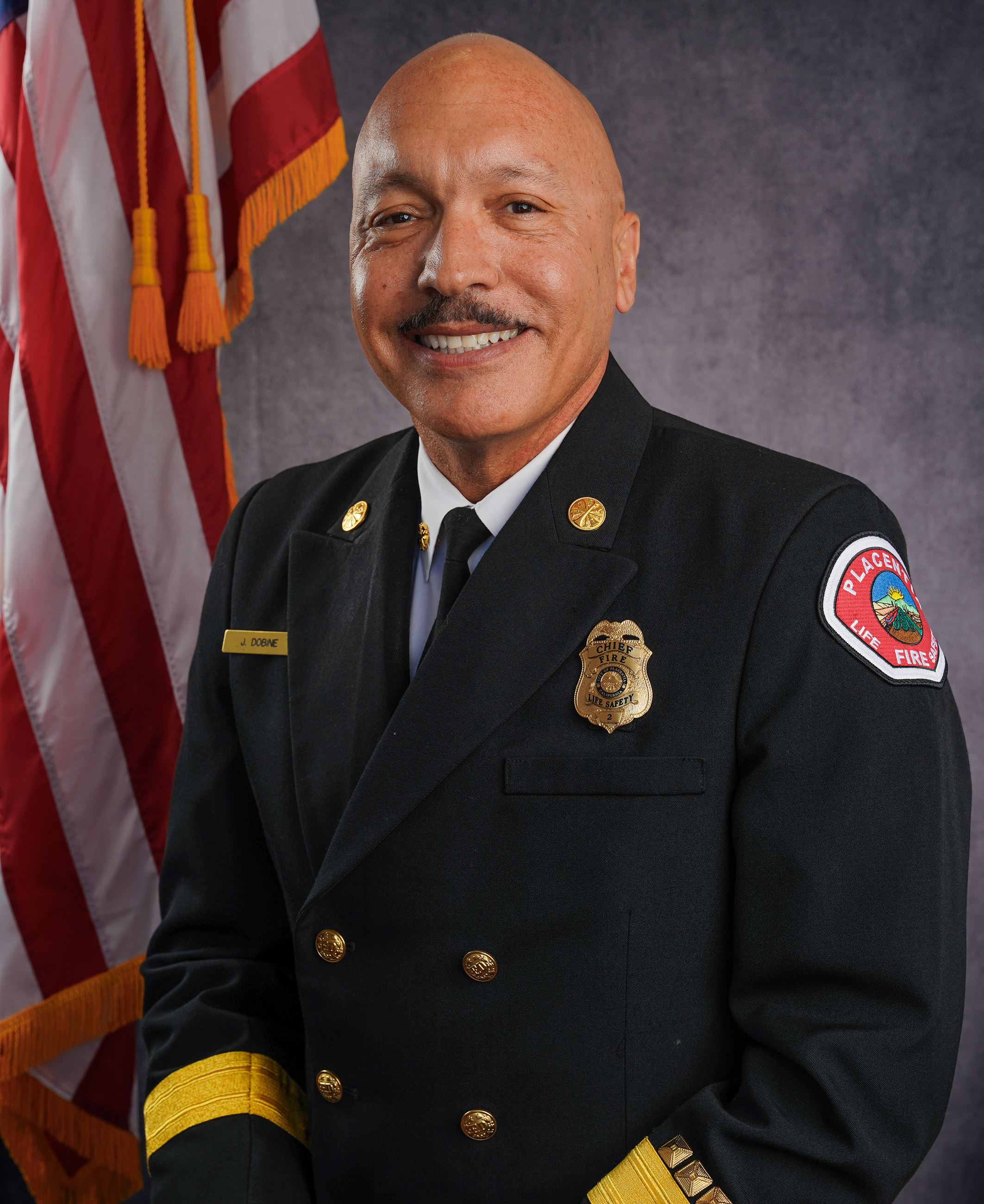Portrait of Placentia Fire Chief Jason Dobine in dress uniform, smiling in front of an American flag