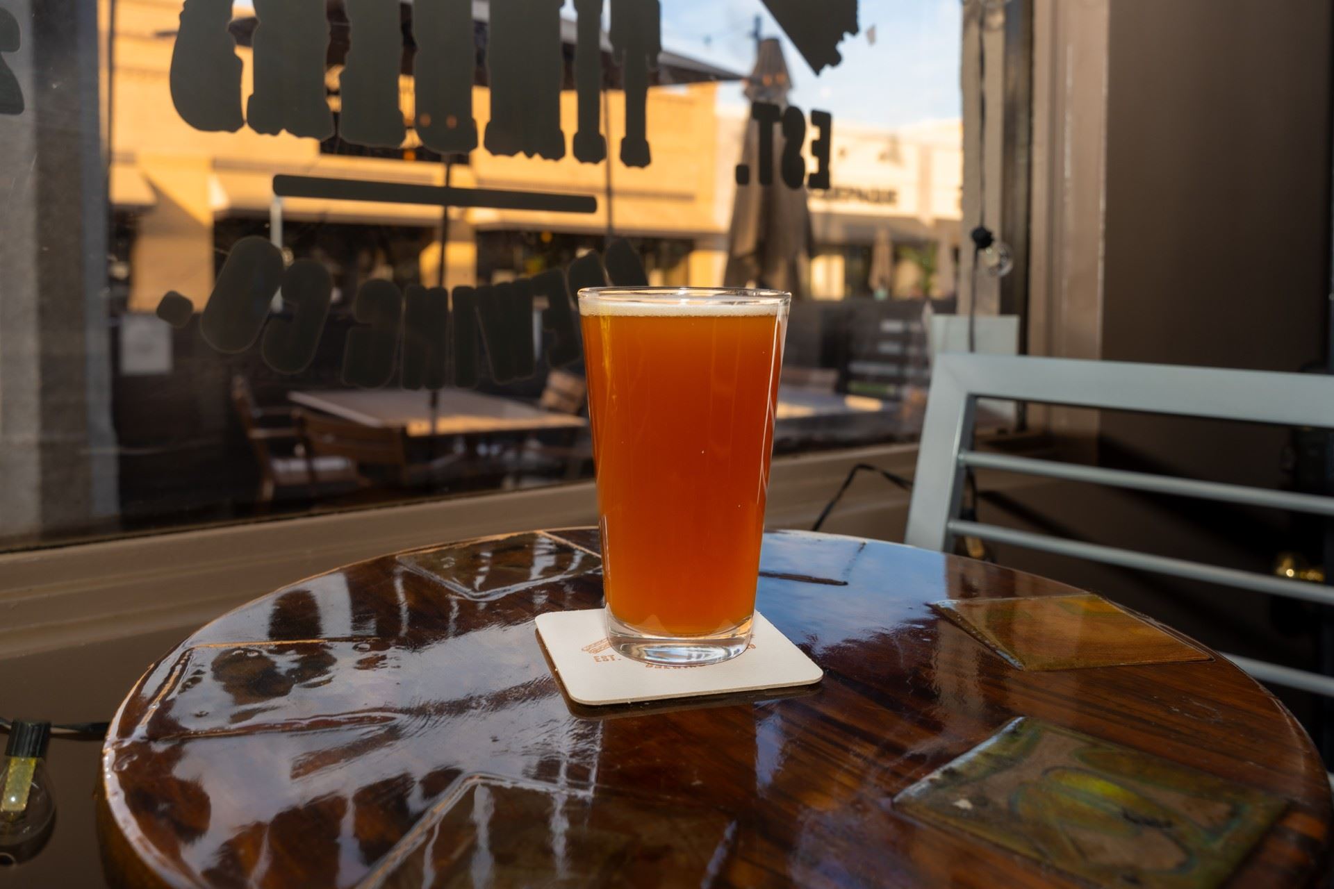 A pint glass filled with amber beer sits on a coaster atop a wooden table inside  a bar with large w…